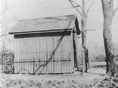 Side view of Fort Conley, a six by eight foot wooden building with a sloped roof. Low metal fencing surrounds it.