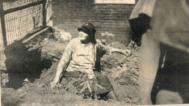 Lyda sits in a ditch dug by workers. A brick building is behind her and the ditch appears to be 2-3 feet deep.