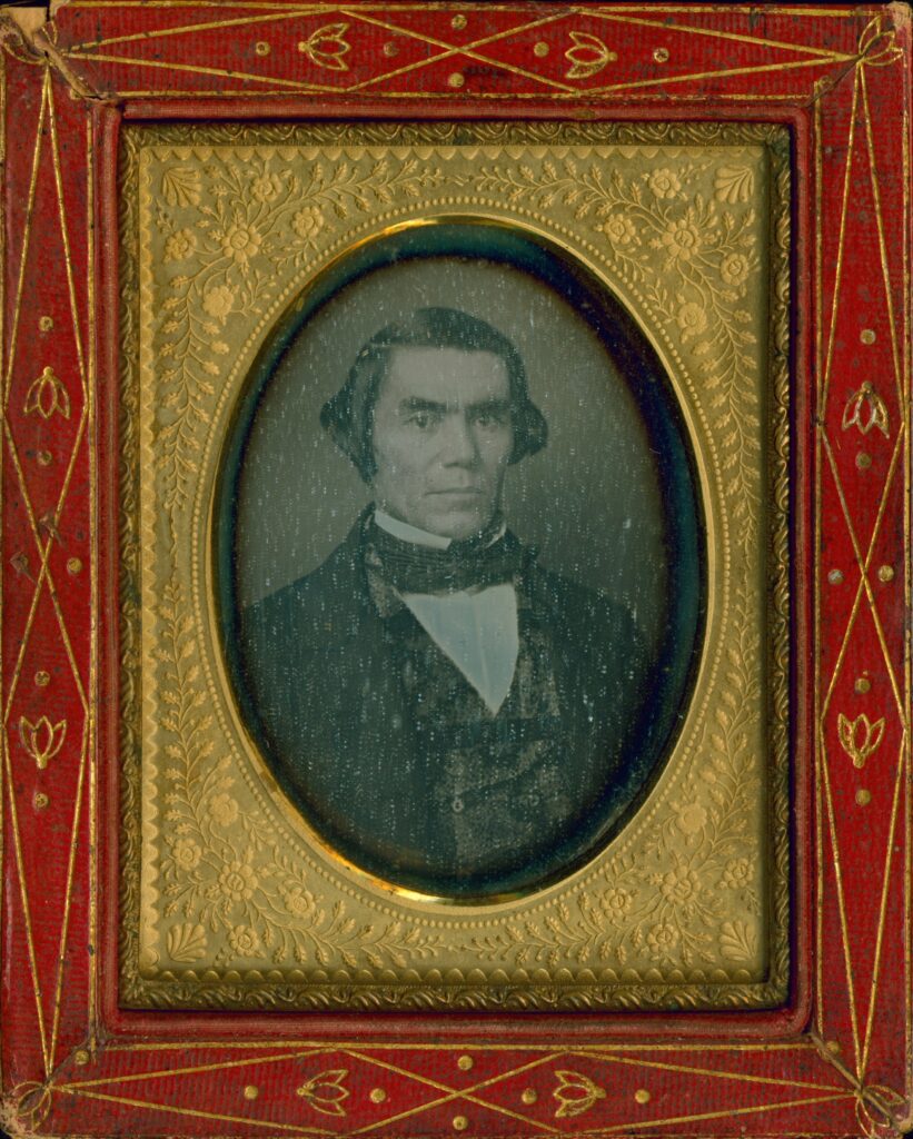 Portrait of Silas Armstrong. Placed in a red and gold frame, he wears a suit from the 1850s.