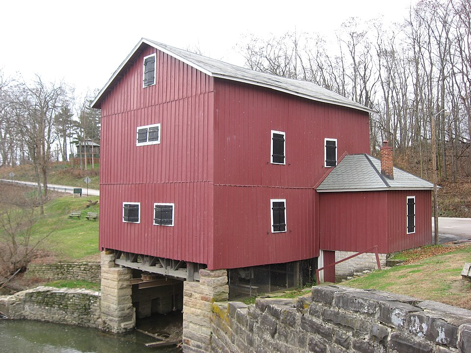 Red metal mill building standing on top of a stone wall that leads to a creek.